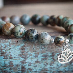 May include: A green and brown stone bracelet with a silver lotus charm. The bracelet is made of round beads and is strung on a silver chain.