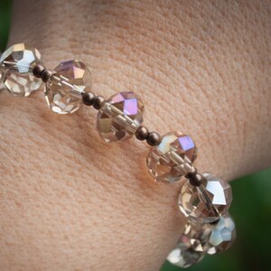 May include: A beaded bracelet featuring faceted, clear, and iridescent crystals. The bracelet is strung on a dark brown cord with small, round beads. The crystals reflect light, creating a sparkling effect. Jewelry.