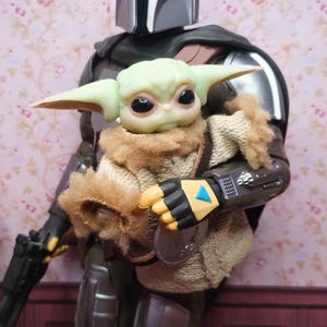 Op de afbeelding: Een gedetailleerd actiefiguur van The Mandalorian die Grogu, ook bekend als Baby Yoda, vasthoudt. De Mandalorian draagt zilveren bepantsering met een zwarte cape, terwijl Grogu in een bruine mantel is gewikkeld. Het figuur staat tegen een bloemenbehang.