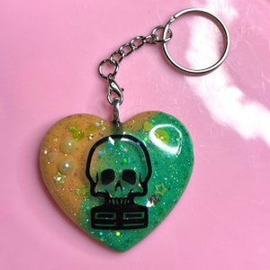 Pode incluir: Um chaveiro de resina em forma de coração verde e laranja com um design de caveira e ossos cruzados pretos. O chaveiro tem glitter embutido na resina.