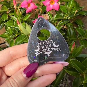 May include: A black and silver holographic planchette with the text "It can't rain all the time" and a crescent moon and star design.