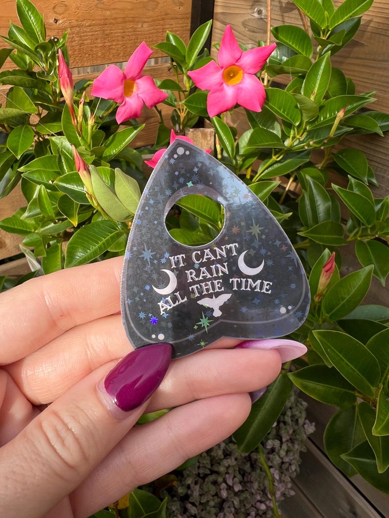 May include: A black and silver holographic planchette with the text "It can't rain all the time" and a crescent moon and star design.