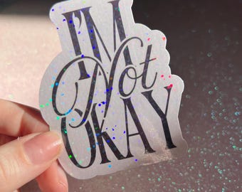 May include: A holographic sticker with the text "I'M Not OKAY" in black. The sticker has a white border. The background is a glittery pink surface. The sticker is held by a hand.