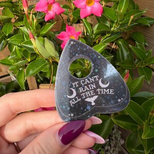 May include: A black and silver glitter-covered  planchette with the text "It can't rain all the time" and a crescent moon and bird design.