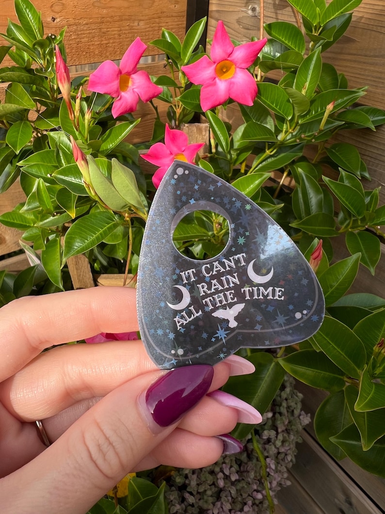 May include: A black and silver glitter-covered  planchette with the text "It can't rain all the time" and a crescent moon and bird design.