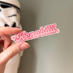 May include: A pink sticker with the word "Kenobiii" in a bubble font, decorated with small flower accents. The sticker is held in front of a blurred white and black figure.