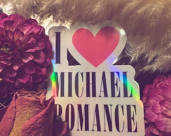 May include: A sticker with the text "I ❤️ MICHAEL ROMANCE" in black capital letters, with a pink heart. The sticker has a holographic border. The sticker is surrounded by dried flowers and fluffy beige grass.