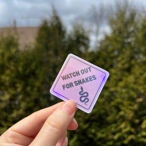 May include: A pink square sticker with a white border and the text "Watch Out For Snakes" in black. A black snake silhouette is below the text.