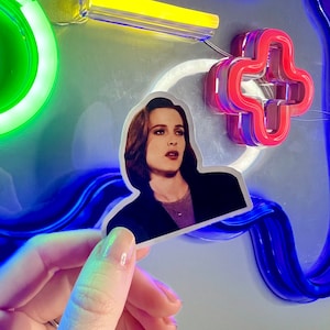 Peut inclure: Un gros plan sur un autocollant représentant une femme aux cheveux brun foncé et une veste noire. L'autocollant est tenu dans une main sur un fond de néons bleus et verts.