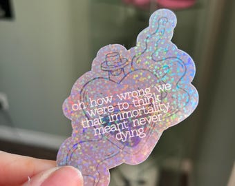May include: A holographic sticker with a heart-shaped design and the text "oh how wrong we were to think that immortality meant never dying." The sticker has a hat-wearing figure and a glittery finish.
