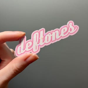 Könnte beinhalten: Rosafarbener Aufkleber mit dem Wort "Deftones" in weißer, kursiv geschriebener Schrift mit rosa Umrandung. Der Aufkleber hat eine glitzernde Oberfläche.