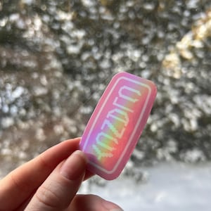 May include: A rectangular sticker with a pink and holographic design. The sticker has a white border and the word "IDZUT" in pixelated rainbow text. The sticker is held in a hand against a blurred outdoor background.
