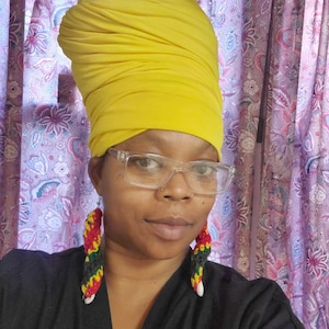 May include: A person wearing a yellow head wrap, clear glasses, and black and red earrings. They are wearing a black shirt with a purple heart badge.