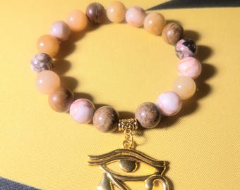 May include: A bracelet made of light brown, pink, and white stones with a gold charm of the Eye of Horus.