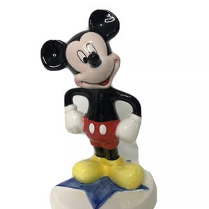 Op de afbeelding: Een keramiek beeldje van Mickey Mouse dat op een blauwe ster staat. Mickey draagt zijn kenmerkende rode broek, gele schoenen en witte handschoenen. Hij heeft een zwart lichaam en een wit gezicht met zwarte ogen en een brede glimlach.