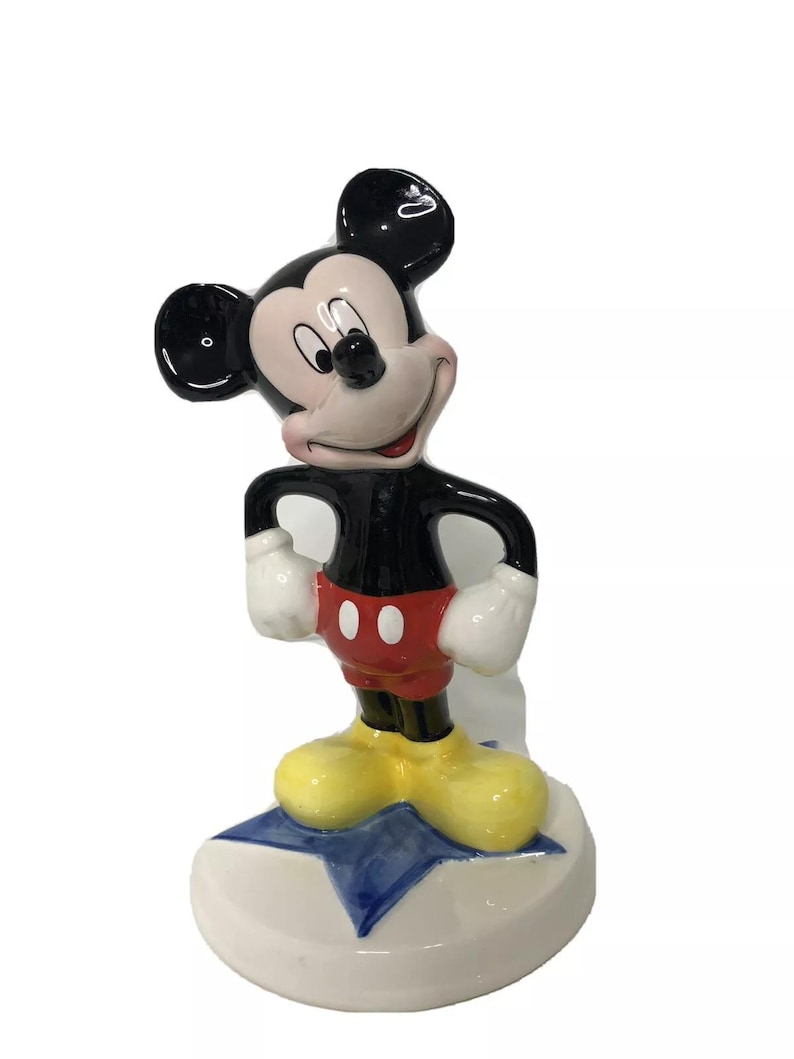 Op de afbeelding: Een keramiek beeldje van Mickey Mouse dat op een blauwe ster staat. Mickey draagt zijn kenmerkende rode broek, gele schoenen en witte handschoenen. Hij heeft een zwart lichaam en een wit gezicht met zwarte ogen en een brede glimlach.