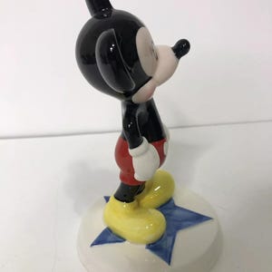 Op de afbeelding: Een keramiek beeldje van Mickey Mouse dat op een blauwe ster staat. Mickey draagt zijn kenmerkende rode broek, gele schoenen en witte handschoenen. Hij kijkt naar rechts.