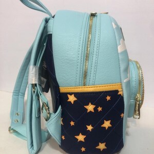 May include: A light blue backpack with gold zippers and straps. The side panel features a navy blue quilted design with yellow stars. The top panel has white clouds. The backpack has a top handle and adjustable straps.