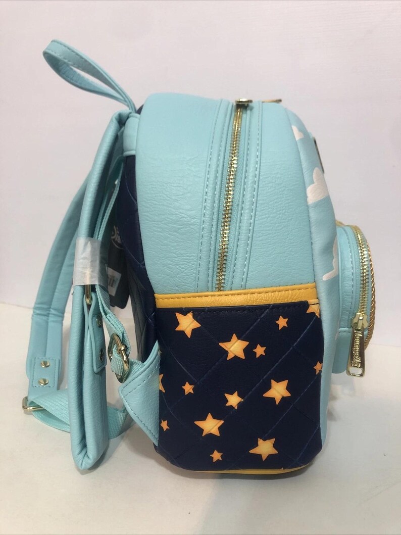 May include: A light blue backpack with gold zippers and straps. The side panel features a navy blue quilted design with yellow stars. The top panel has white clouds. The backpack has a top handle and adjustable straps.
