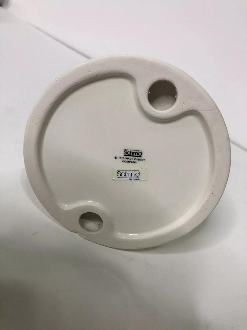 Op de afbeelding: Witte keramische figuurbasis met een gat om op te hangen. De basis heeft de tekst "Schmid" en "The Walt Disney Company" erop gedrukt.