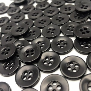 May include: A large group of black plastic buttons with four holes each. The buttons are arranged in a random pattern on a white surface.