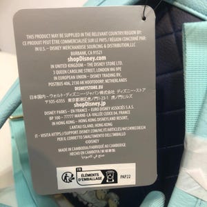 May include: A product tag with text in multiple languages, including English, French, and Japanese. The tag lists the product's origin, Disney's contact information, and recycling symbols. The tag is attached to a light blue and navy blue item.