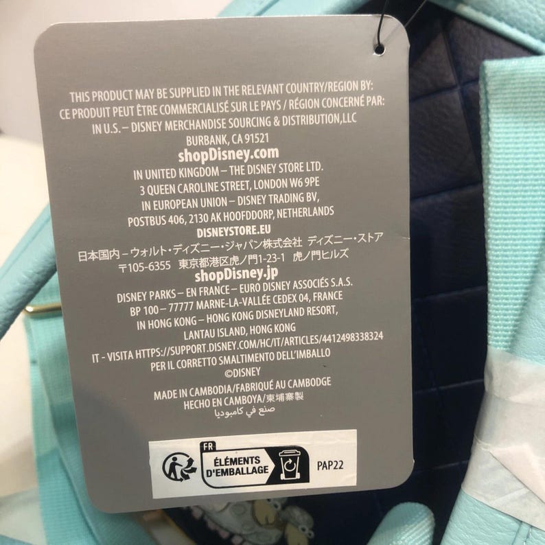 May include: A product tag with text in multiple languages, including English, French, and Japanese. The tag lists the product's origin, Disney's contact information, and recycling symbols. The tag is attached to a light blue and navy blue item.