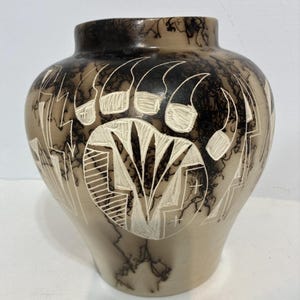 May include: A round ceramic vase with a smooth, light beige surface featuring dark, wispy black smoke-like patterns. The exterior is decorated with deeply etched white carvings of a large bear paw print and geometric lightning bolt shapes.