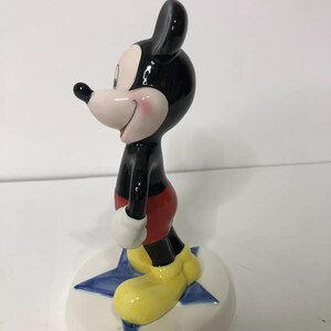 Op de afbeelding: Een keramiek beeldje van Mickey Mouse dat op een blauwe ster staat. Mickey draagt zijn kenmerkende rode broek, gele schoenen en zwart-witte handschoenen. Hij heeft een grote glimlach op zijn gezicht.
