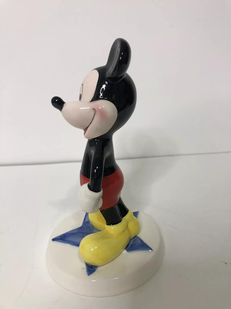 Op de afbeelding: Een keramiek beeldje van Mickey Mouse dat op een blauwe ster staat. Mickey draagt zijn kenmerkende rode broek, gele schoenen en zwart-witte handschoenen. Hij heeft een grote glimlach op zijn gezicht.