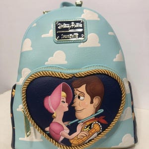 May include: A light blue backpack featuring a cloud pattern and a heart-shaped design with Woody and Bo Peep from Toy Story. The backpack has a gold-colored zipper and trim, and a metal plaque that reads "Disney Parks Loungefly."