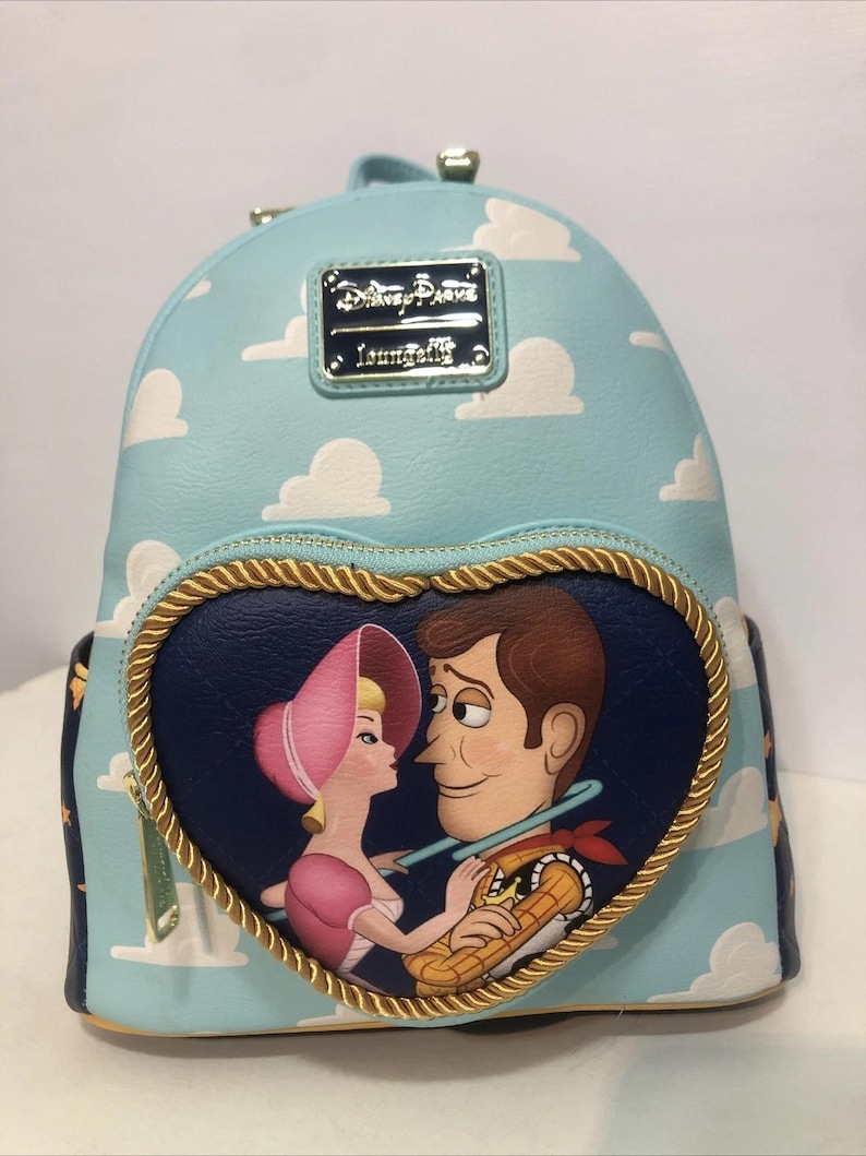 May include: A light blue backpack featuring a cloud pattern and a heart-shaped design with Woody and Bo Peep from Toy Story. The backpack has a gold-colored zipper and trim, and a metal plaque that reads "Disney Parks Loungefly."