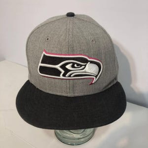 May include: Gray and black baseball cap with a pink and white Seattle Seahawks logo. The logo features a stylized hawk with a fierce expression.