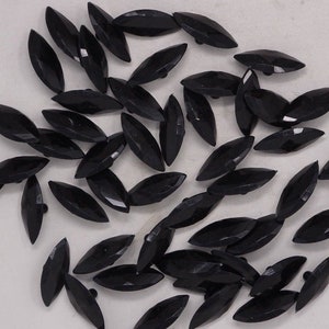 May include: A collection of black, faceted, marquise-shaped buttons. These glossy buttons have a pointed oval shape and are scattered across a light-colored surface. The buttons are likely made of plastic or glass and are suitable for sewing projects.