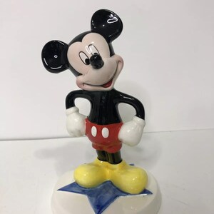 Op de afbeelding: Een keramiek beeldje van Mickey Mouse dat op een blauwe ster staat. Mickey draagt zijn kenmerkende rode broek met twee zwarte knopen, gele schoenen en witte handschoenen. Hij heeft een zwart lichaam en hoofd met grote zwarte oren en een wit gezicht.