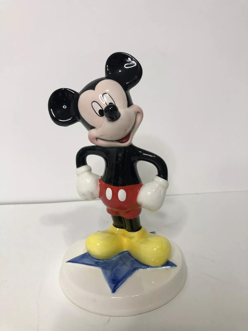 Op de afbeelding: Een keramiek beeldje van Mickey Mouse dat op een blauwe ster staat. Mickey draagt zijn kenmerkende rode broek met twee zwarte knopen, gele schoenen en witte handschoenen. Hij heeft een zwart lichaam en hoofd met grote zwarte oren en een wit gezicht.
