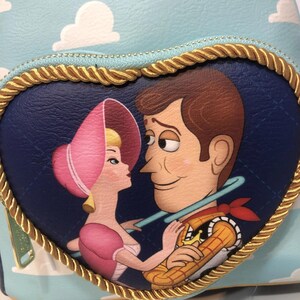 May include: A heart-shaped purse featuring a cartoon image of Bo Peep and Woody from Toy Story. The image is set against a blue background with white clouds. The purse has a gold rope trim and a light blue zipper.