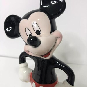 Op de afbeelding: Een keramiek beeldje van Mickey Mouse. Mickey staat met zijn armen gekruist en draagt zijn kenmerkende rode broek met witte knopen. Hij heeft zwarte schoenen en witte handschoenen. Mickey glimlacht en heeft een zwarte neus en grote zwarte oren.