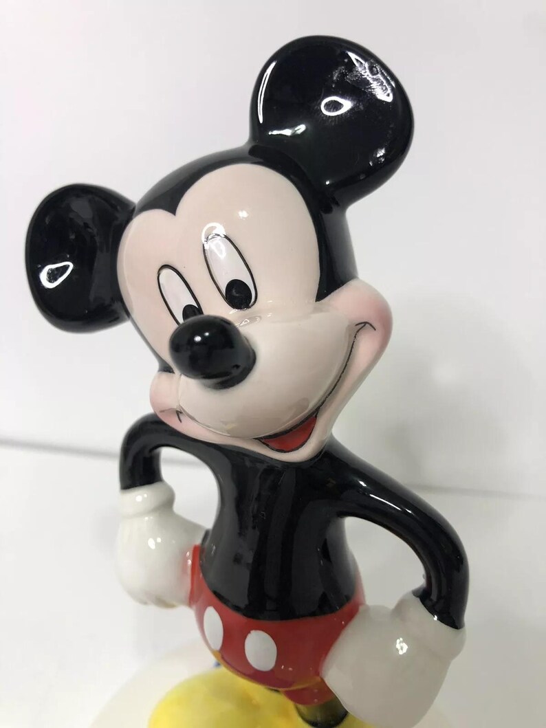 Op de afbeelding: Een keramiek beeldje van Mickey Mouse. Mickey staat met zijn armen gekruist en draagt zijn kenmerkende rode broek met witte knopen. Hij heeft zwarte schoenen en witte handschoenen. Mickey glimlacht en heeft een zwarte neus en grote zwarte oren.