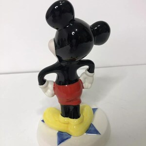 Op de afbeelding: Een keramiek beeldje van Mickey Mouse dat op een blauwe en witte stervormige basis staat. Mickey draagt zijn kenmerkende rode broek en gele schoenen.