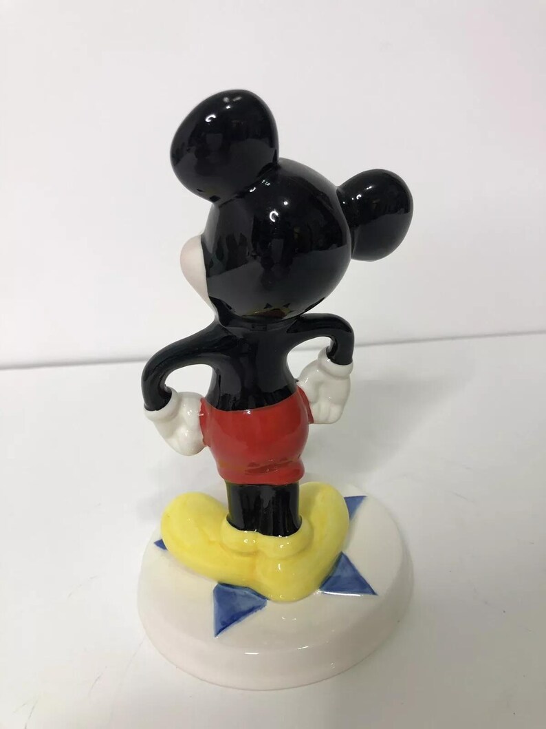 Op de afbeelding: Een keramiek beeldje van Mickey Mouse dat op een blauwe en witte stervormige basis staat. Mickey draagt zijn kenmerkende rode broek en gele schoenen.