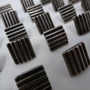 May include: A collection of silver-toned metal buttons with a ridged design. The buttons are square shaped and have a slightly raised edge.