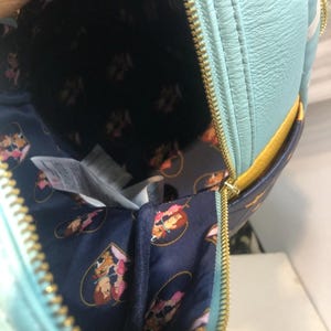 May include: A light blue backpack with a gold zipper, open to reveal a navy blue interior with a repeating cartoon character pattern. The backpack has a yellow accent and a white stripe.
