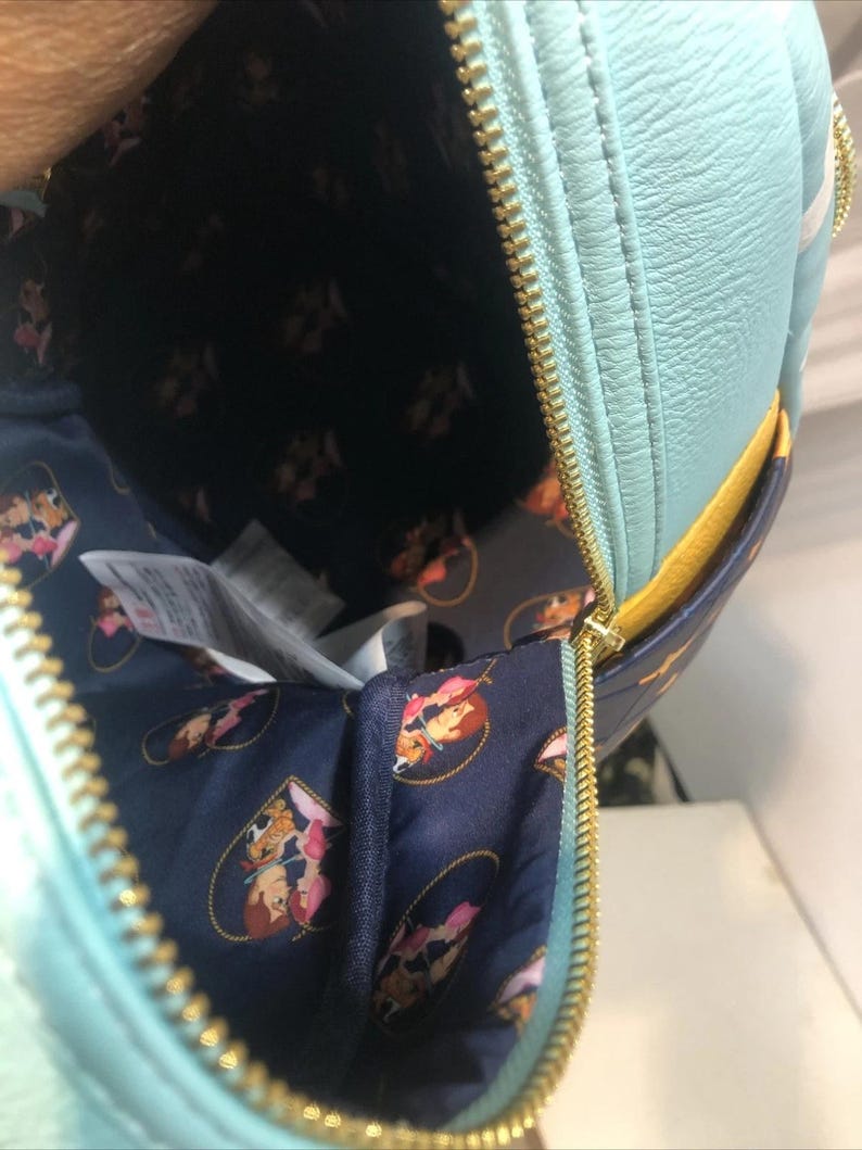May include: A light blue backpack with a gold zipper, open to reveal a navy blue interior with a repeating cartoon character pattern. The backpack has a yellow accent and a white stripe.