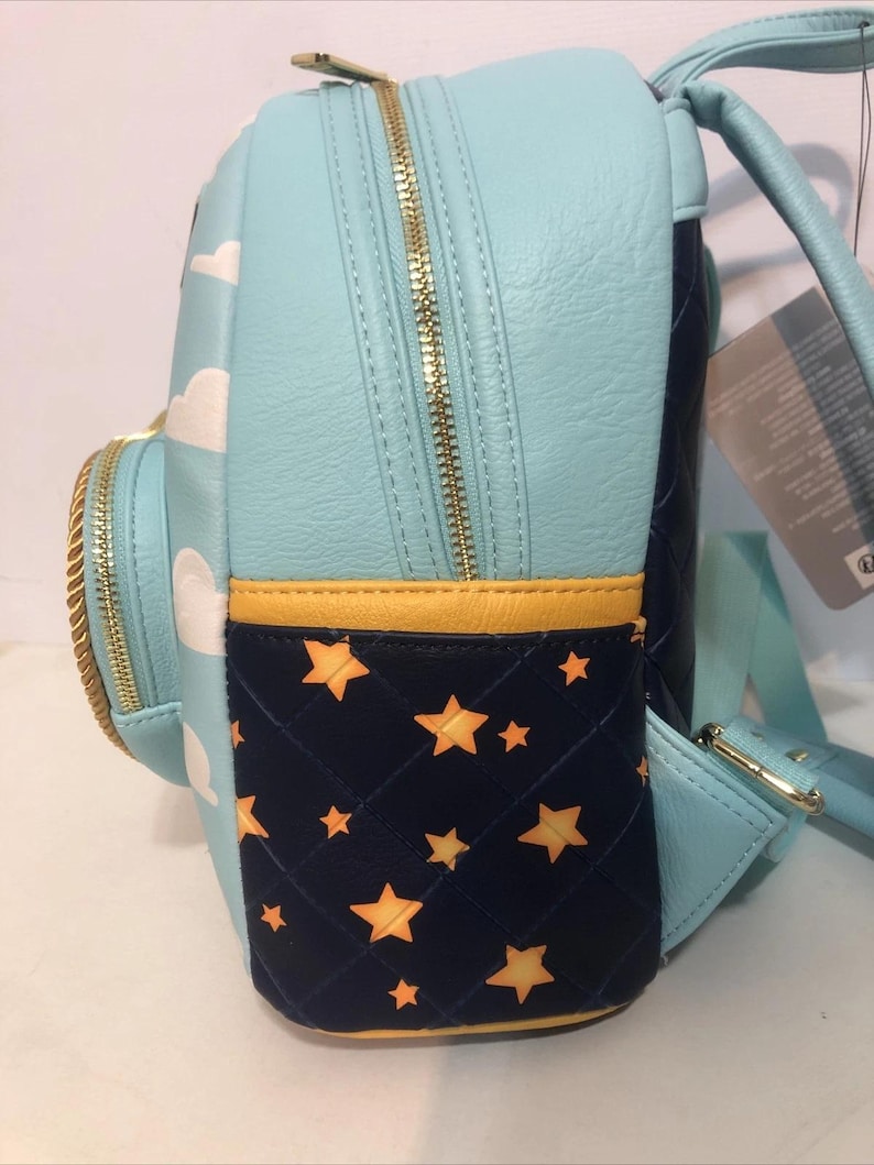 May include: A light blue backpack with white cloud designs and gold zippers. The side pocket is navy blue with a quilted pattern and gold stars. The backpack has a yellow trim.