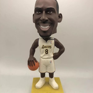 May include: A bobblehead figurine of Kobe Bryant wearing a white Los Angeles Lakers jersey with the number 8 on the front. He is holding a basketball in his left hand and has a big smile on his face. The base of the figurine is yellow and says "Kobe Bryant" in black letters.