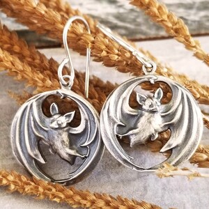 May include: A pair of silver earrings featuring a detailed bat design. Each earring has a circular shape with a cutout bat silhouette in the center. The bat has spread wings and a detailed face.