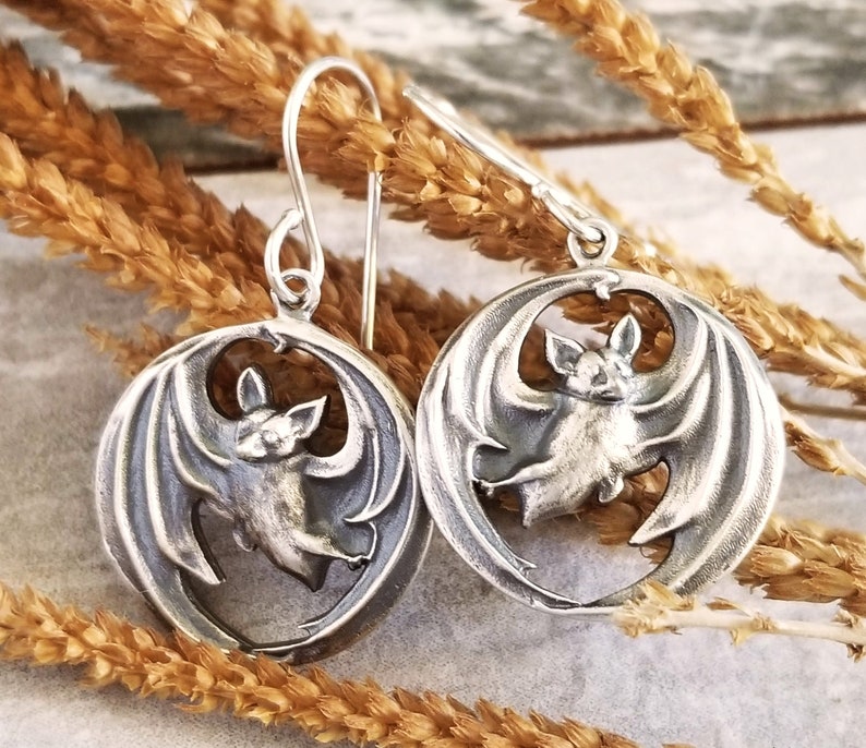 May include: A pair of silver earrings featuring a detailed bat design. Each earring has a circular shape with a cutout bat silhouette in the center. The bat has spread wings and a detailed face.