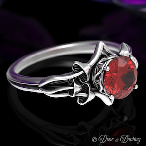 Puede incluir: Un anillo de plata con una piedra preciosa roja engastada en un diseño con forma de hongo. El anillo es intrincado y detallado, con un delicado patrón de enredadera.