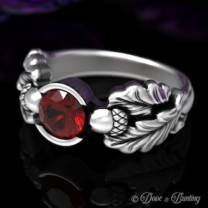 May include: A silver ring with a red gemstone set in a round bezel. The ring is decorated with two oak leaves and acorns on either side of the stone.